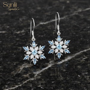 May include: A pair of silver snowflake earrings with blue and white gemstones. Each earring features a central square blue stone surrounded by white and blue marquise-cut stones, and a silver hook.