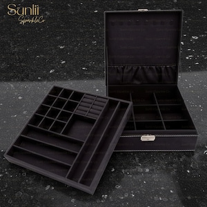 以下が含まれることがあります： 複数のコンパートメントと取り外し可能なトレイを備えた黒いジュエリーボックス。ボックスは開いており、裏地付きの内部とジュエリーを整理するためのさまざまなセクションが表示されます。トレイには、リングやその他の小さなアイテム用のスロットがあります。ボックスは暗い表面にあります。