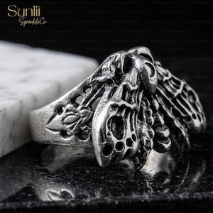 May include: A silver-toned ring with an intricate moth design. The ring features detailed wings and a textured surface, showcasing a gothic or alternative aesthetic. The ring is displayed on a dark, reflective surface.