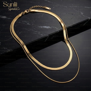 May include: A layered gold-toned necklace with a herringbone chain and a delicate, thin chain. The necklace is displayed on a dark surface. The brand name "Sunlii Sparkle Co" is visible in the upper left corner.