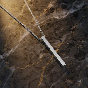 May include: A silver bar pendant necklace with a fine chain. The pendant is a simple, rectangular shape, hanging vertically. The necklace is displayed on a dark marble surface. The brand name "Sunlii SparkleCo" is in the top left corner.