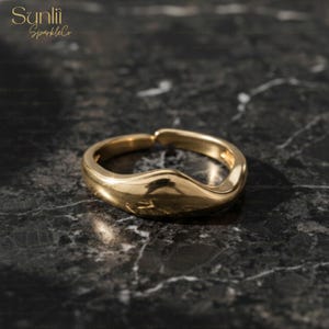 May include: A gold-colored ring with a wavy design. The ring is made of a shiny, reflective metal and sits on a dark, marbled surface. The ring's design features a smooth, curved shape, adding a touch of elegance.