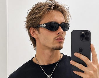 Peut inclure: Lunettes de soleil noires avec des accents latéraux argentés, un collier à chaîne à billes argenté avec un pendentif pointu, et un t-shirt noir à manches longues avec des motifs graphiques tribaux blancs et le texte "MOBTDMIDE 2BEEDCOBE".