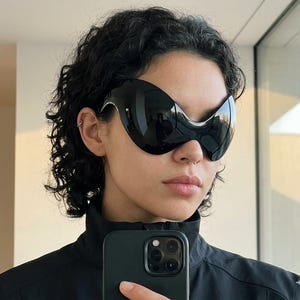 May include: Oversized black sunglasses with a continuous, smooth, curved frame that covers the upper face. The lenses are dark and reflective, featuring a singular, fluid shape that connects across the bridge of the nose without a visible frame edge.
