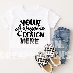 以下が含まれることがあります： 白いTシャツに黒い文字で「YOUR Awesome DESIGN HERE」と書いてあります。Tシャツは白い表面に置かれており、黒と白のチェック柄のスリッポンシューズと青いジーンズが置いてあります。