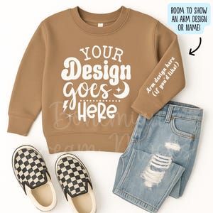 May include: A tan sweatshirt with the text "YOUR Design goes Here" in white, paired with light blue denim jeans and black and white checkered slip-on shoes. The sweatshirt has space for an arm design or name.