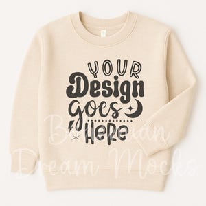 Może przedstawiać: Kremowa bluza z napisem "YOUR Design goes Here" w czarnym, retro kroju pisma. Projekt zawiera gwiazdę, błyskawicę i półksiężyc. Bluza ma okrągły dekolt i długie rękawy.