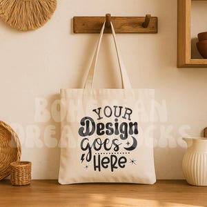 Puede incluir: Un bolso tote de color crema con el texto "YOUR Design goes Here" en escritura negra. El bolso tiene asas largas de color blanquecino y se muestra sobre una superficie de madera. El fondo incluye elementos decorativos y un colgador de pared de madera.