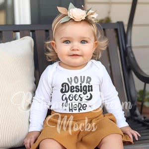 May include: A baby wearing a white long-sleeved onesie with the text "YOUR Design goes HERE" in black. The baby is also wearing a mustard-colored corduroy skirt and a floral headband. The baby is sitting on a black bench.