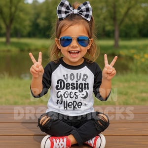 Op de afbeelding: Een zwart-wit baseball-T-shirt voor kinderen met de tekst "YOUR Design goes HERE". Het kind draagt een zwarte ripped jeans, rode sneakers en een zwart-wit geruite strik. Het kind draagt een blauwe spiegelende zonnebril.