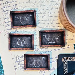 Puede incluir: Cuatro pequeñas pizarras rectangulares con marcos de madera oscura. Cada pizarra presenta un dibujo con tiza blanca de un caldero con brazos y piernas. Las pizarras están sobre un fondo de papel vintage.