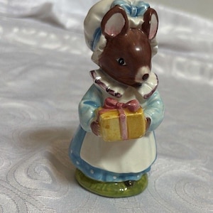 Puede incluir: Figura de cerámica de un ratón con un vestido azul, un delantal blanco y un gorro, sosteniendo una caja de regalo amarilla atada con una cinta rosa. El ratón tiene pelaje marrón y orejas rosas. La figura está sobre una base verde.