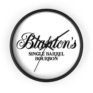 May include: A round wall clock with a black frame and a white face. The clock face features the words "Blanton's SINGLE BARREL BOURBON" in black script. The clock hands are black.