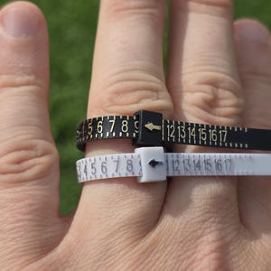May include: Two plastic ring sizers, one black and one white, each featuring a sliding buckle with an arrow indicator and printed numerical markings ranging from 1 to 17, designed to measure finger circumference for determining ring size.