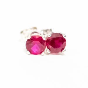 May include: A pair of stud earrings featuring round, vibrant red gemstones set in a silver metal. The earrings are displayed against a white background, highlighting the color and shape of the stones. The gemstones have a polished finish.