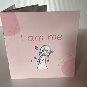 May include: A pink card with the text "I am me" and an illustration of a smiling person surrounded by hearts. The card has a light pink background with decorative star and oval shapes.