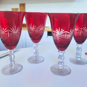 May include: Four ruby red wine glasses with clear stems and bases. Each glass features a decorative etched design near the top. The glasses are arranged on a white surface, suggesting a formal setting or special occasion.