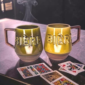 Peut inclure: Deux chopes à bière en céramique, une verte olive et une jaune, avec le mot "BIERE" en blanc. Elles ont des anses marron et sont posées sur une surface en bois avec des cartes à jouer.