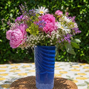 Peut inclure: Un arrangement floral vibrant composé de grandes pivoines roses, de marguerites blanches, de statice violet et de petites fleurs de remplissage vertes. Le bouquet est présenté dans un vase en céramique bleu profond et brillant.