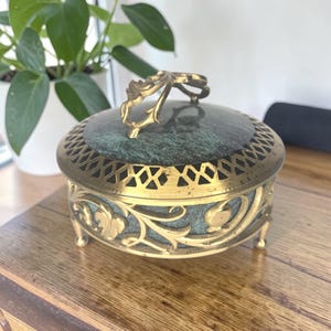 May include: An antique jewelry box with a green marble top and ornate gold-colored metal detailing. The box features a decorative handle and intricate floral patterns around the sides. It rests on small, curved feet.