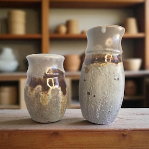 Peut inclure: Deux vases en céramique faits à la main, d'esthétique rustique. Les vases sont dans des tons de gris et de brun, avec une surface texturée. Le plus petit vase mesure 10 cm de haut, et le plus grand vase mesure 15 cm de haut. Ils sont exposés sur une surface en bois.