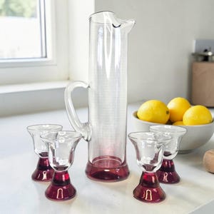 Peut inclure: Une grande carafe en verre transparent avec une anse incurvée et une base de couleur magenta profond, accompagnée de quatre petits verres assortis, chacun doté d'un bol supérieur transparent et d'une base magenta solide, posés sur une surface.