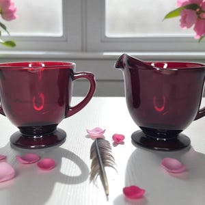 Peut inclure: Un ensemble de service à crème et sucrier en verre rouge rubis. Le pot à crème a un bec et une poignée, tandis que le sucrier a deux poignées. Des pétales de fleurs roses et une plume sont éparpillés autour de l'ensemble.