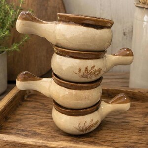 Peut inclure: Une pile de quatre bols à soupe en céramique avec poignées, présentant un corps couleur crème et un bord émaillé marron foncé. Deux des bols sont décorés d'un motif d'épis de blé marron sur le côté, tandis que les autres sont unis.
