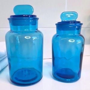 May include: Two matching blue glass apothecary jars with lids. The jars are cylindrical with a rounded base and a matching lid with a small handle. The jars are a vibrant blue colour and are likely used for storage or decoration.