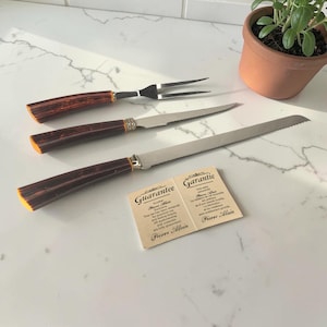 Peut inclure: Un ensemble de couverts Pierre Alain comprenant une fourchette à découper, un couteau dentelé et un couteau à pain. Les manches sont bruns et les lames en acier inoxydable. Une carte de garantie est présente.