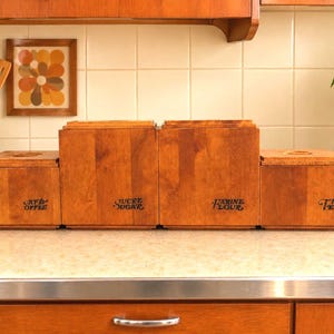 May include: A set of four rectangular wooden kitchen canisters with lids, arranged in a stepped formation. The canisters are labelled in black text with both French and English: Café Coffee, Sucre Sugar, Farine Flour, and The Tea.