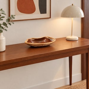 Puede incluir: Un cuenco de cerámica ovalado con un interior liso de color marrón oscuro y un borde texturizado con motas beige y blancas. El cuenco tiene una forma orgánica irregular y se asienta sobre una superficie plana, mostrando el contraste entre el centro oscuro y el borde claro.