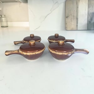 May include: A set of four small brown ceramic bowls with matching lids and side handles. Each bowl features a glossy dark brown finish on the lid and base, accented by a textured, lighter tan band around the rim where the lid meets the bowl.