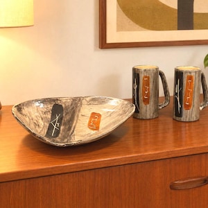 May include: A ceramic bowl and two matching mugs featuring a grey and white marbled finish. Each piece is decorated with a vertical black rectangular accent containing white line art and a separate orange rectangular accent with white line art.