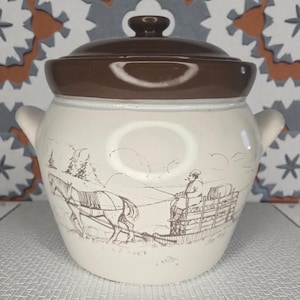 May include: A vintage, cream-coloured ceramic crock pot with a brown lid and handles. The crock pot features a sepia-toned illustration of a horse-drawn sleigh in a winter scene. The lid has a small, round handle.