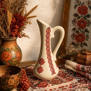 May include: A cream-colored ceramic pitcher featuring a tall, slender neck, a curved handle, and a decorative vertical band of red and black geometric embroidery-style patterns, complemented by two red floral motifs on the lower body.