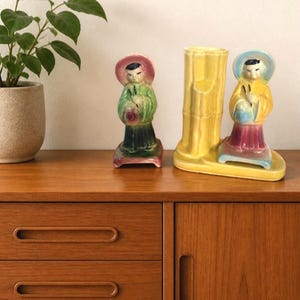 Peut inclure: Deux figurines en céramique et un vase sont posés sur une surface en bois. Les figurines, vêtues de costumes traditionnels, sont vertes et jaunes. Le vase jaune présente un motif de bambou. Une plante en pot est visible en arrière-plan.