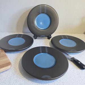 Peut inclure: Un ensemble de quatre assiettes modernes en céramique. Chaque assiette a un anneau extérieur gris foncé et un centre bleu clair. Les assiettes sont disposées sur une surface blanche, avec une planche à découper en bois et un couteau.
