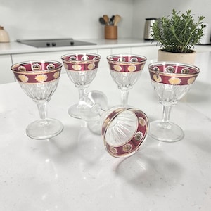 Peut inclure: Ensemble de cinq verres à vin en verre transparent avec une bande décorative rouge et dorée sur le bord. Les gobelets ont un design classique avec une base à pied et un bol à motifs. Un gobelet est couché sur le côté.