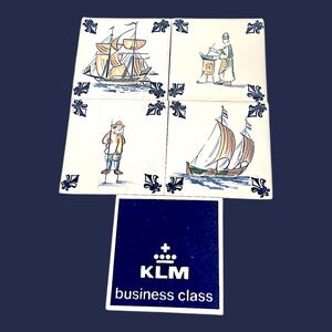 May include: Four square ceramic tiles with nautical scenes and a KLM business class logo. The tiles depict a sailboat, a person working, a person on stilts, and two sailboats. The tiles have a white background with blue and yellow accents.