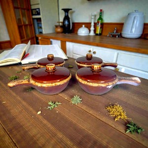 May include: Four brown ceramic soup bowls with lids and handles are displayed on a wooden table. The bowls have a glossy finish and a two-tone design, with a lighter rim. The lids have a decorative knob. Herbs and a cookbook are also on the table.