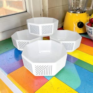 Peut inclure: Quatre bols de service blancs, octogonaux, avec un motif de rayures diagonales grises. Les bols sont disposés sur une surface colorée à motifs arc-en-ciel. Un blender jaune et un bol de fruits sont en arrière-plan.