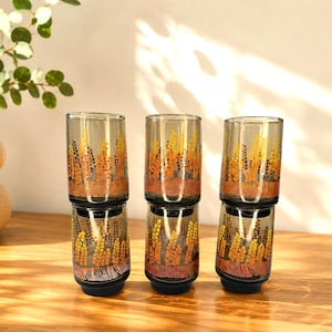Peut inclure: Trois verres cylindriques en verre gris translucide. Chaque verre présente un motif répétitif de formes florales verticales jaunes et orange sur l'extérieur, avec une base gris foncé solide au bas de chaque récipient.