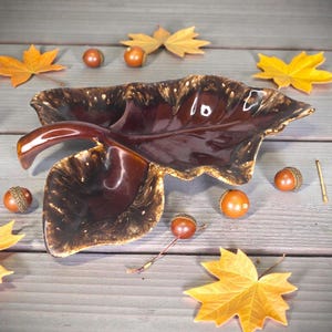 Könnte beinhalten: Eine braune, keramikförmige Servierschale in Blattform mit glänzendem Finish. Die Schale ist von gelben Ahornblättern und Eicheln umgeben, wodurch ein Herbstthema entsteht. Die Schale hat einen einzigartigen Griff und einen strukturierten Rand.