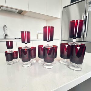 May include: A set of ruby red stemmed glasses. The glasses feature a cylindrical bowl and a clear, circular base. The glasses are displayed on a white surface, with a kitchen and appliances visible in the background.