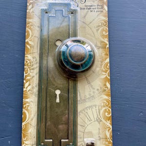 May include: A Graphic 45 Geometric Metal Door Plate and Knob set in a two-piece package. The hardware features an aged bronze finish with a teal blue circular accent on the knob. The rectangular backplate includes a keyhole and mounting holes.