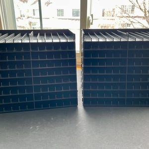 Op de afbeelding: Twee zwarte pennen organizers met meerdere sleuven. Elke organizer heeft een rechthoekige vorm met een reeks verticale verdelers, waardoor individuele compartimenten ontstaan. De organizers zijn gemaakt van een donker materiaal en zijn ontworpen om pennen, potloden of andere schrijfinstrumenten in op te bergen.