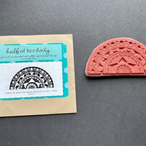 May include: A red rubber stamp with a half-circle design featuring intricate floral and geometric patterns. The stamp is next to a package with the text "half of herdoily" and a matching design. The stamp is for crafts and scrapbooking.