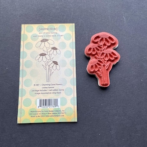 May include: A red rubber stamp of cone flowers next to a package with instructions and a flower illustration. The package includes the text "Charming Cone Flowers" and "Lindsey Spencer". The stamp is mounted on cling foam.