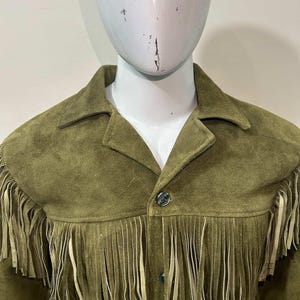 May include: Olive green suede jacket with fringe detailing across the chest and shoulders. The jacket has a collared neckline and button-down front. The fringe is a lighter shade of tan. The jacket is displayed on a mannequin.